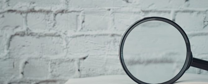 Close-up of a magnifying glass resting on an open notebook with a blurred white brick wall in the background