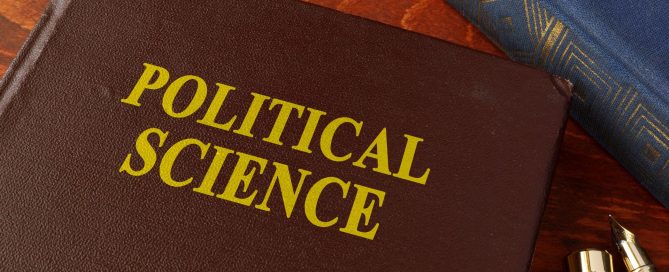 Brown leather book cover with large gold letters reading 'POLITICAL SCIENCE' on a wooden desk, beside a blue fabric and gold-tipped pens.