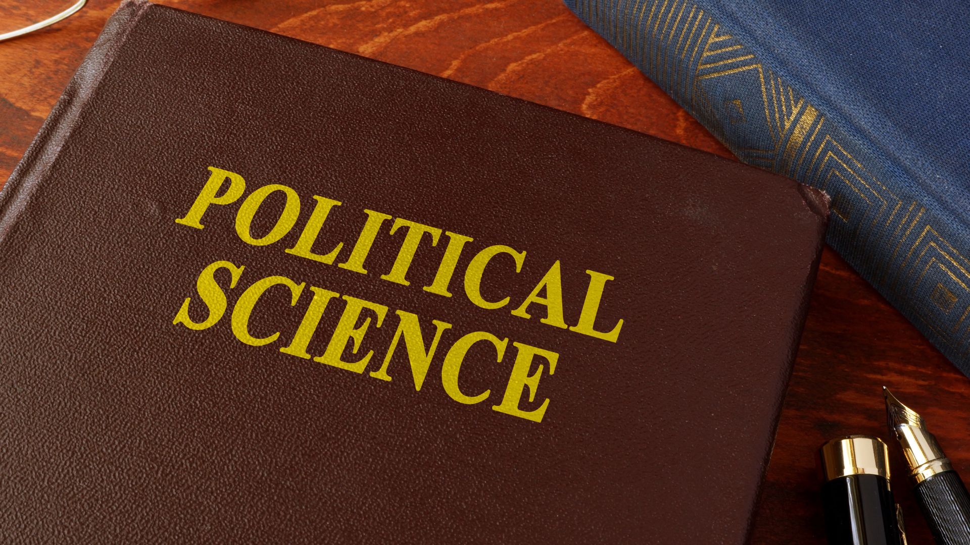 Brown leather book cover with large gold letters reading 'POLITICAL SCIENCE' on a wooden desk, beside a blue fabric and gold-tipped pens.