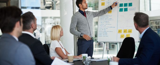 Presenter in a gray cardigan explains a workflow on a whe whiteboard with sticky notes to a seated team in a modern conference room.